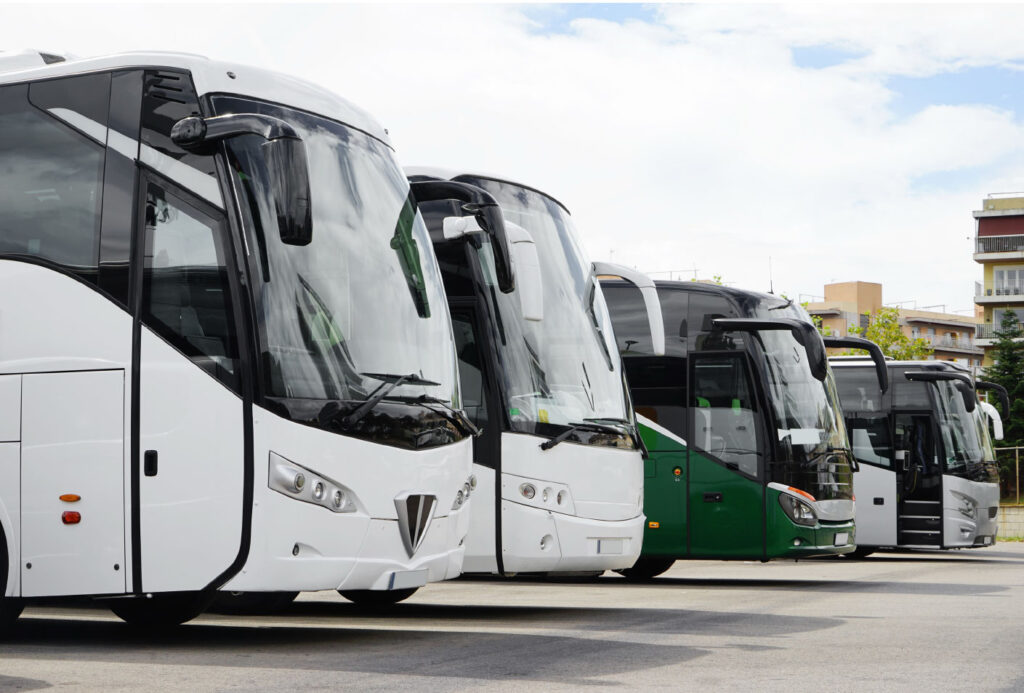 Several modern coach buses, mostly white with one green, are parked in a row near an industrial project site services lot on a cloudy day.