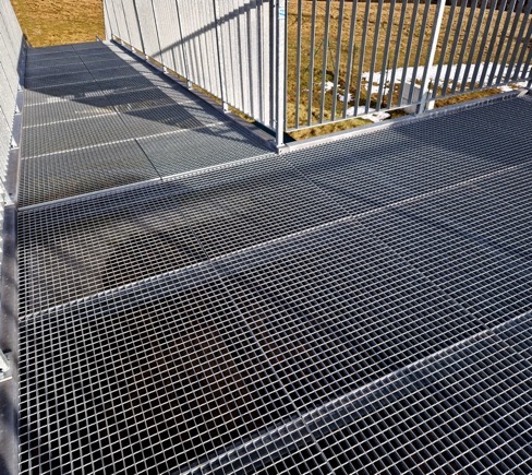 A metal grate walkway with vertical railings, typical of industrial project site services, stands outdoors on a sunny day with grass in the background.