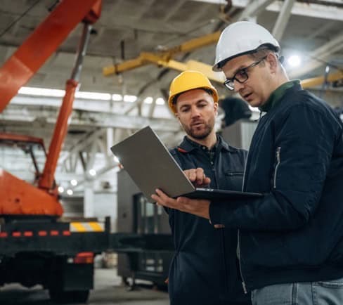 Two men in hard hats stand at an industrial project site, discussing something on a laptop—experts in industrial contractor services.